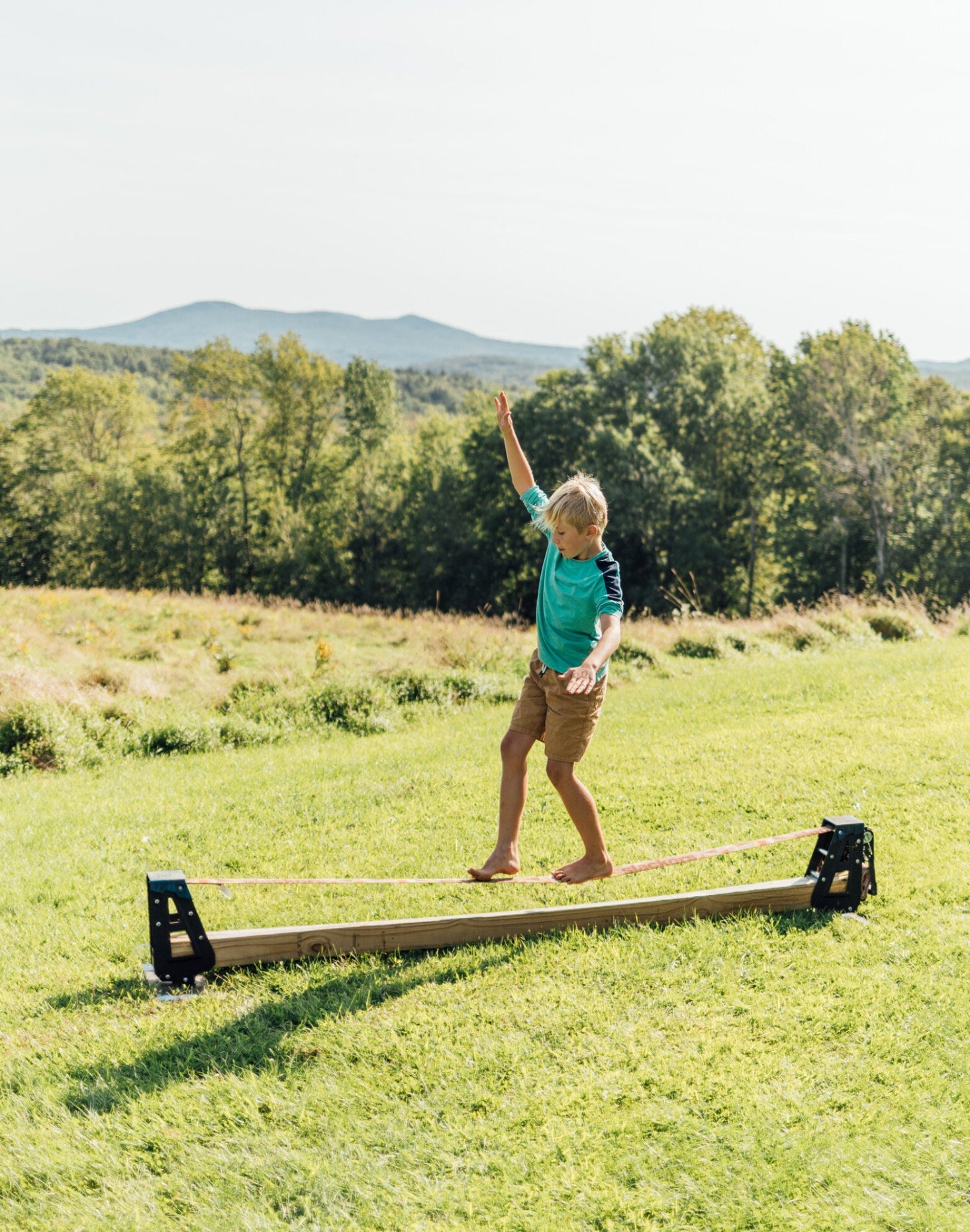 Person standing on a slackline balance beam in a grassy field with trees and mountains in the background