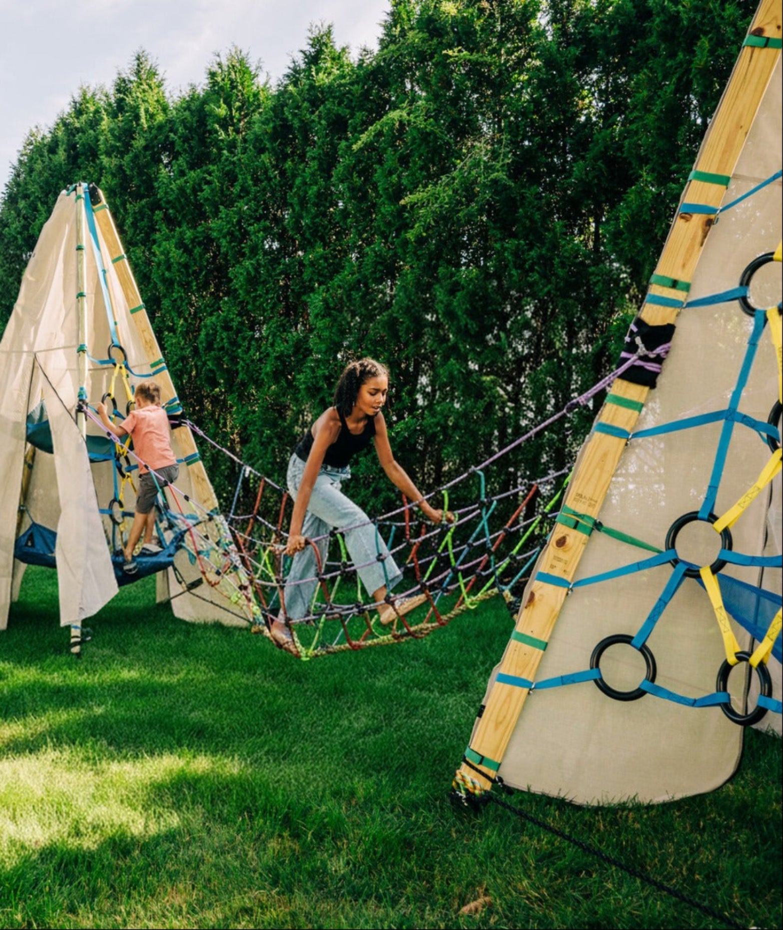 Two children playing on a rope net between two teepees in a grassy outdoor area.