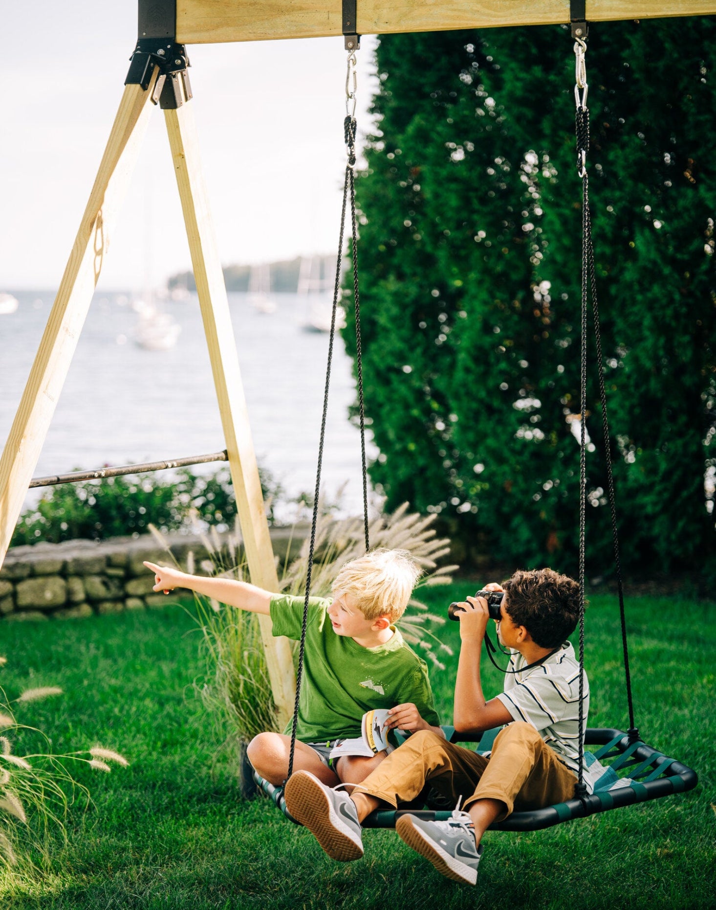 Two children sitting on a modern hexagon-shaped swing with adjustable ropes in an outdoor setting, with one child pointing at something in the distance.