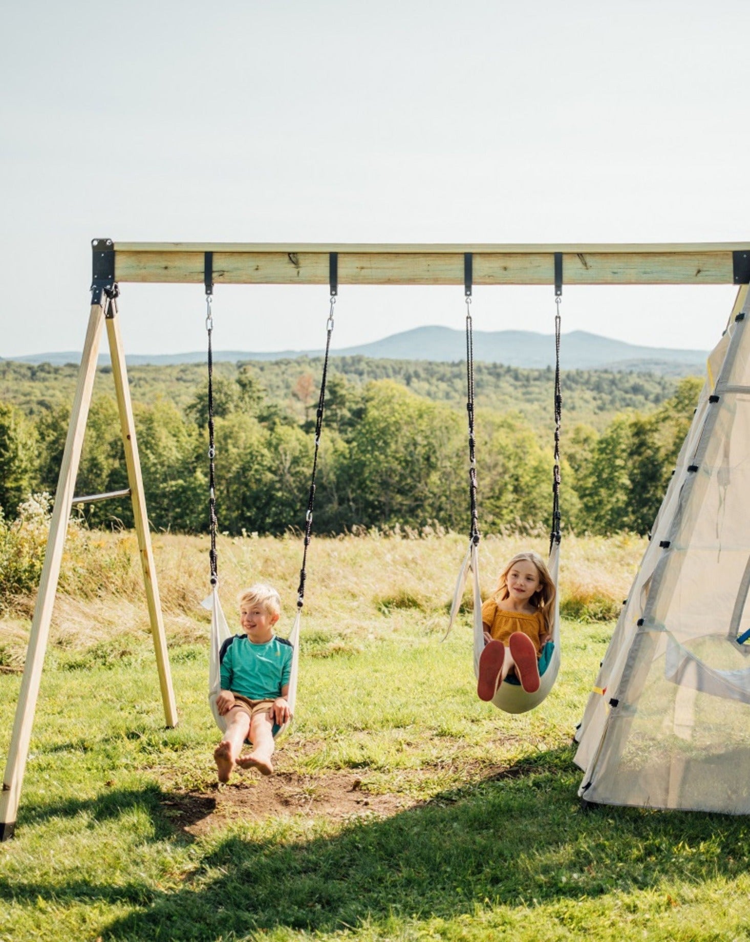 Two children swinging on a two-person swing set in an outdoor grassy area with a white teepee tent beside them.