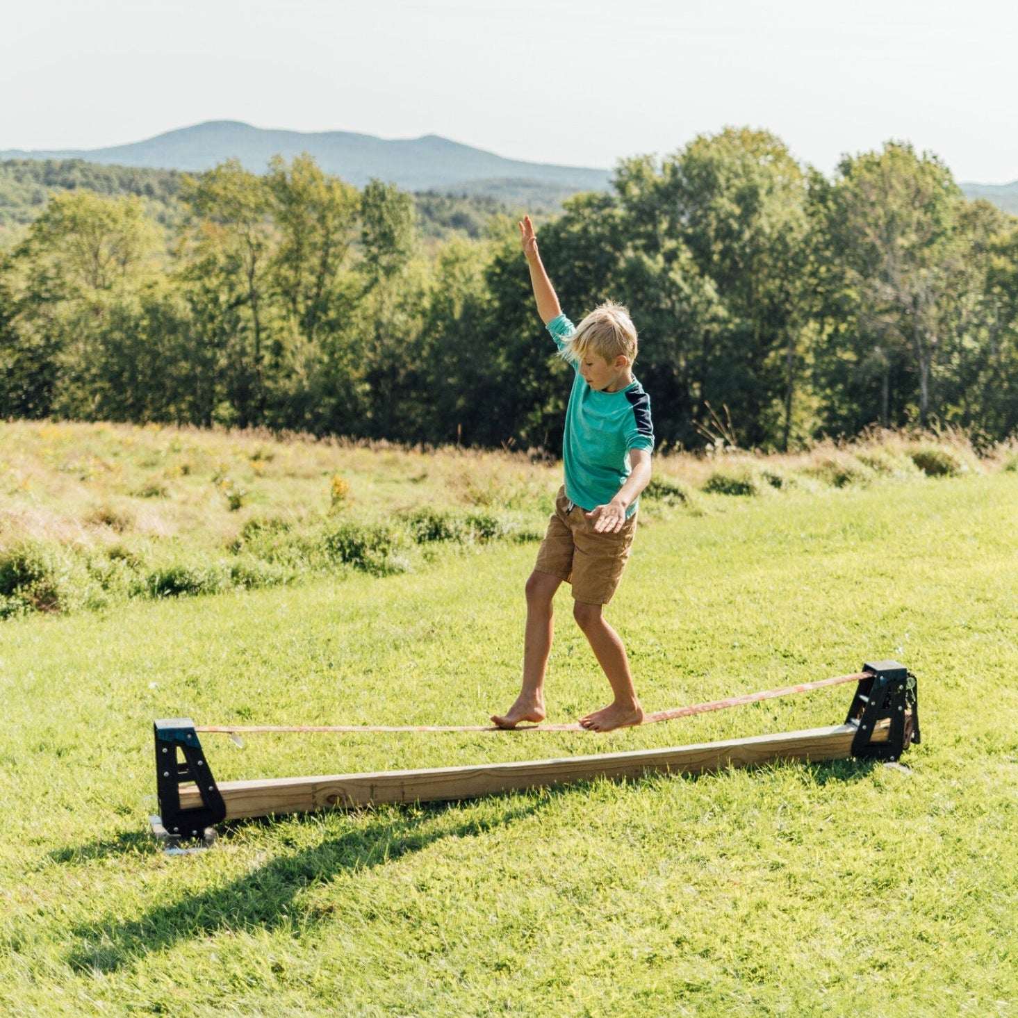 Person standing on a slackline balance beam in a grassy field with trees and mountains in the background