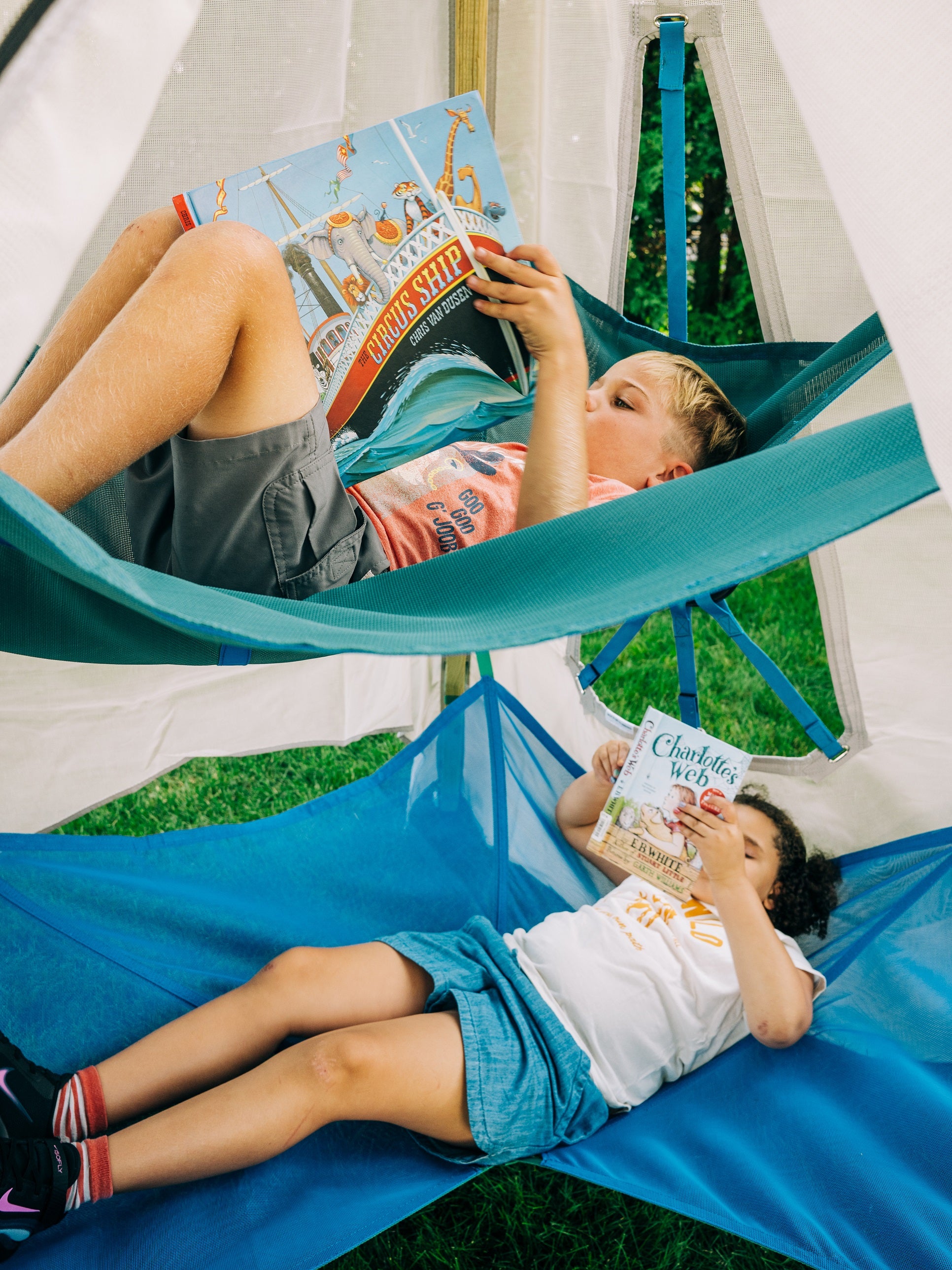 Two children reading books in a hammock outdoors.
