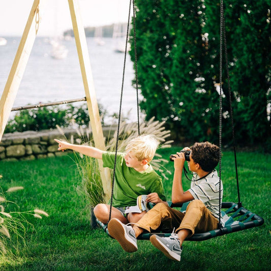 Two children sitting on a modern hexagon-shaped swing with adjustable ropes in an outdoor setting, with one child pointing at something in the distance.
