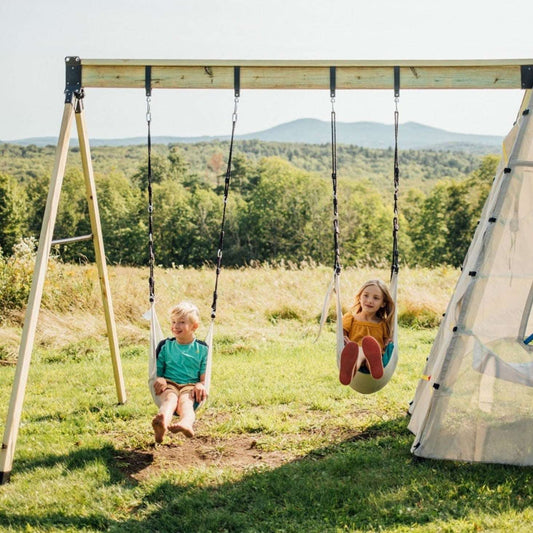 Two children swinging on a two-person swing set in an outdoor grassy area with a white teepee tent beside them.