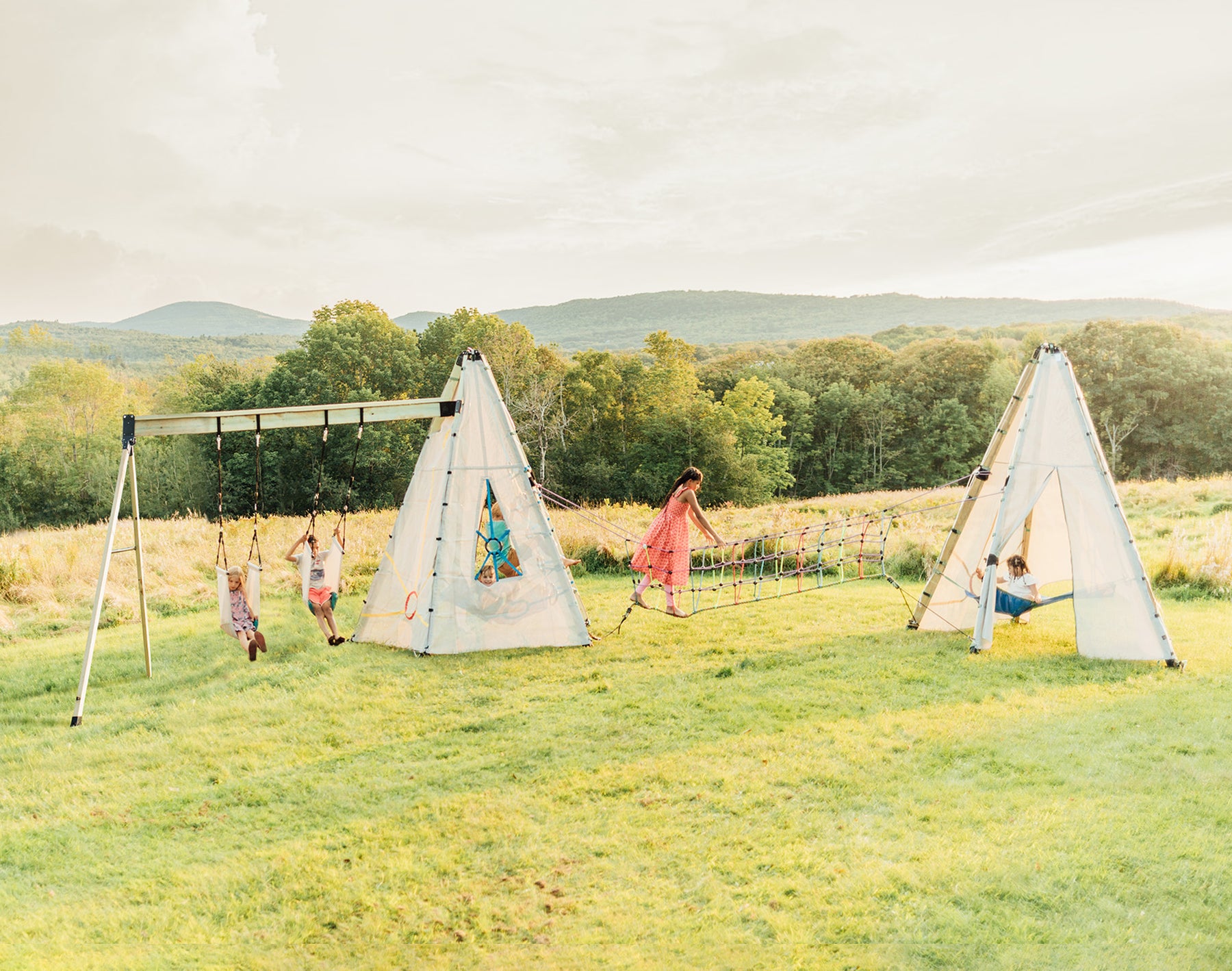 Children play on swings and climbing structures in a grassy field with trees in the background.