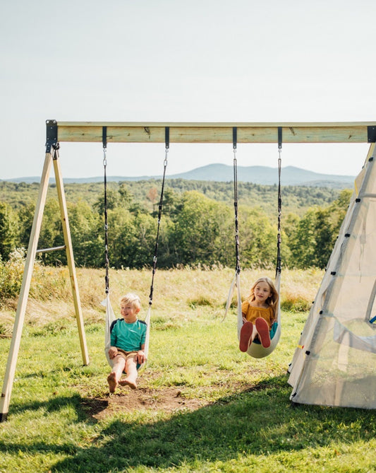 Two children swinging on a two-person swing set in an outdoor grassy area with a white teepee tent beside them.
