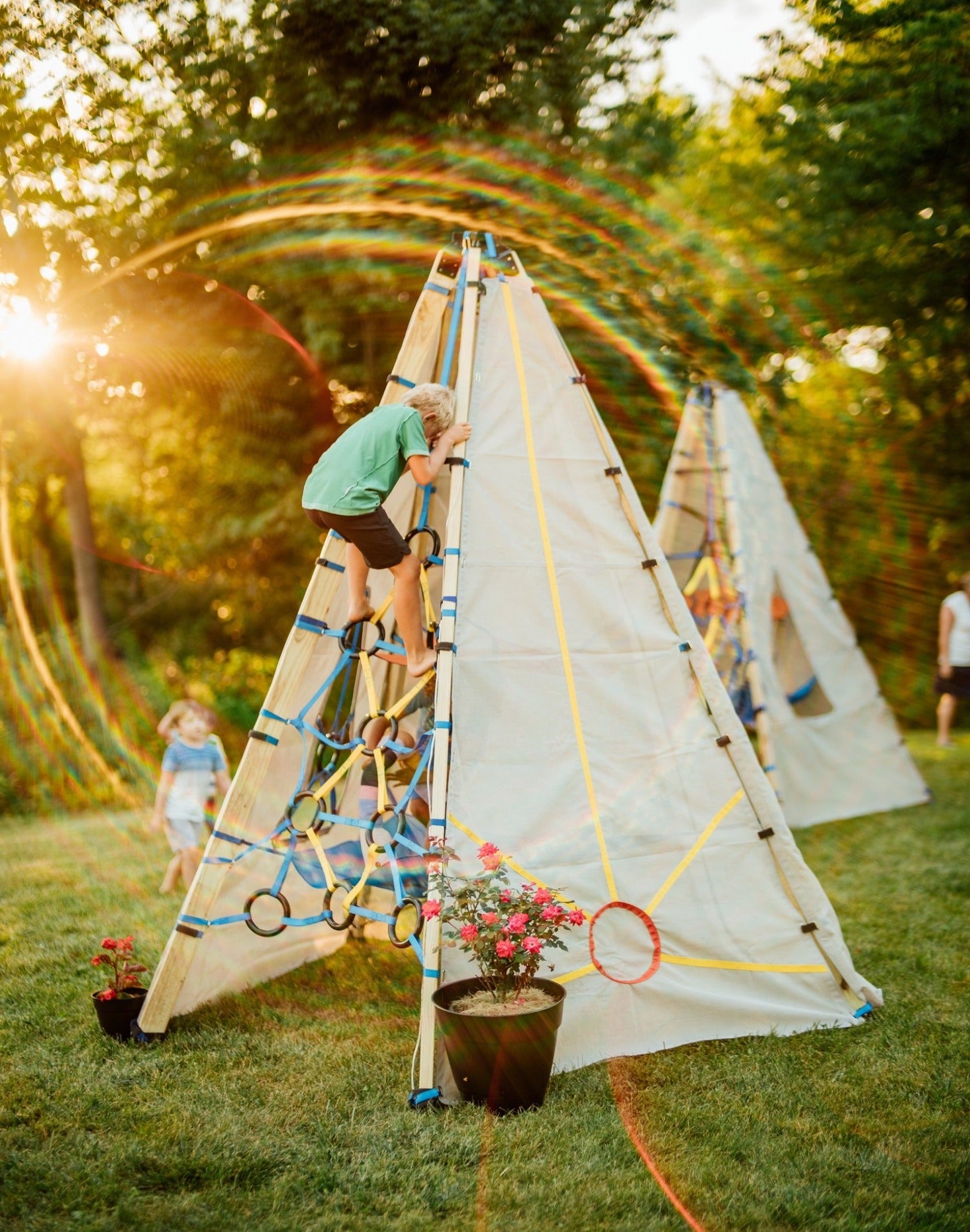 Children climbing a triangular play structure in a sunlit garden setting.