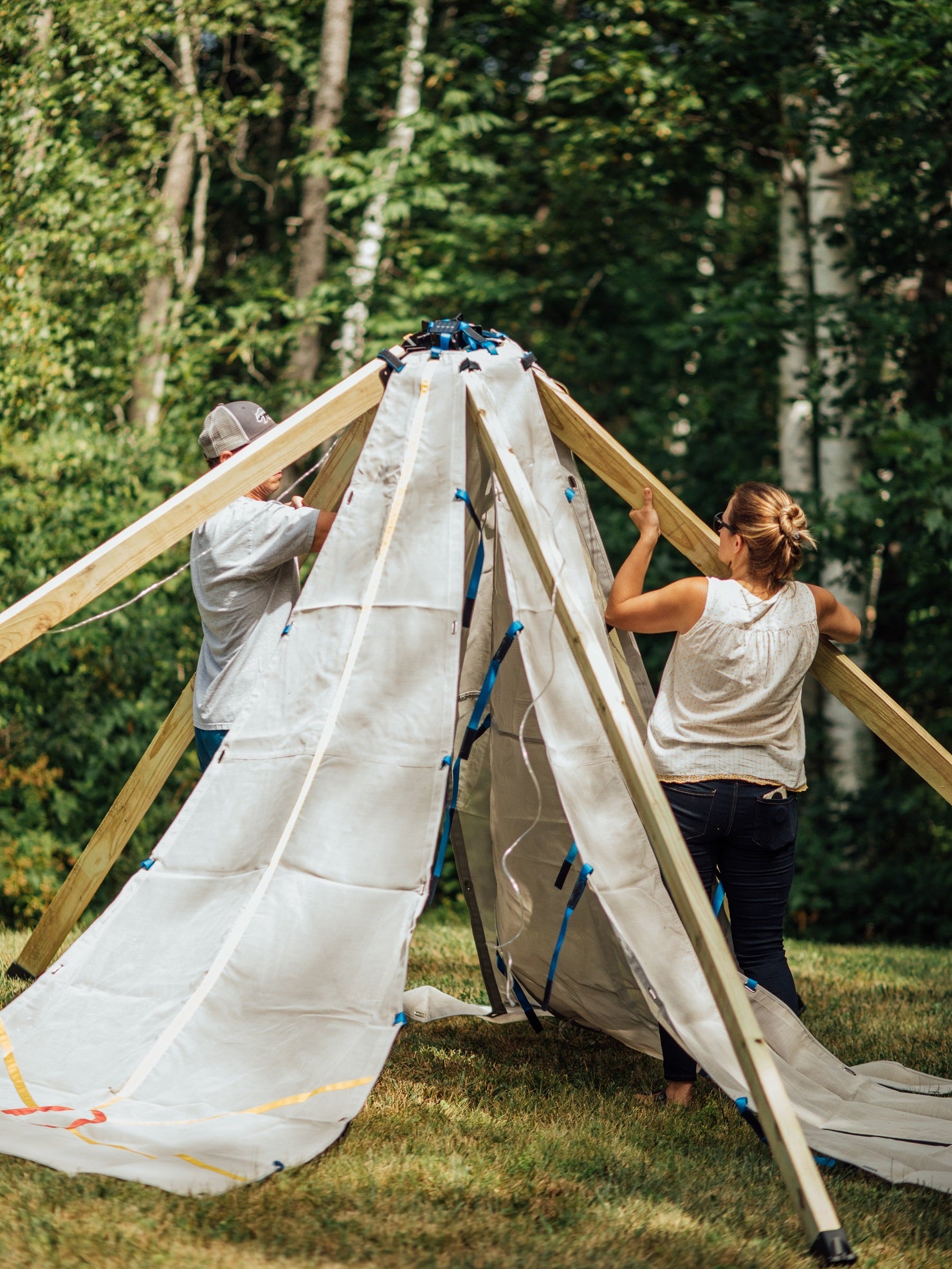 Two people assembling a large tent outdoors in a wooded area.